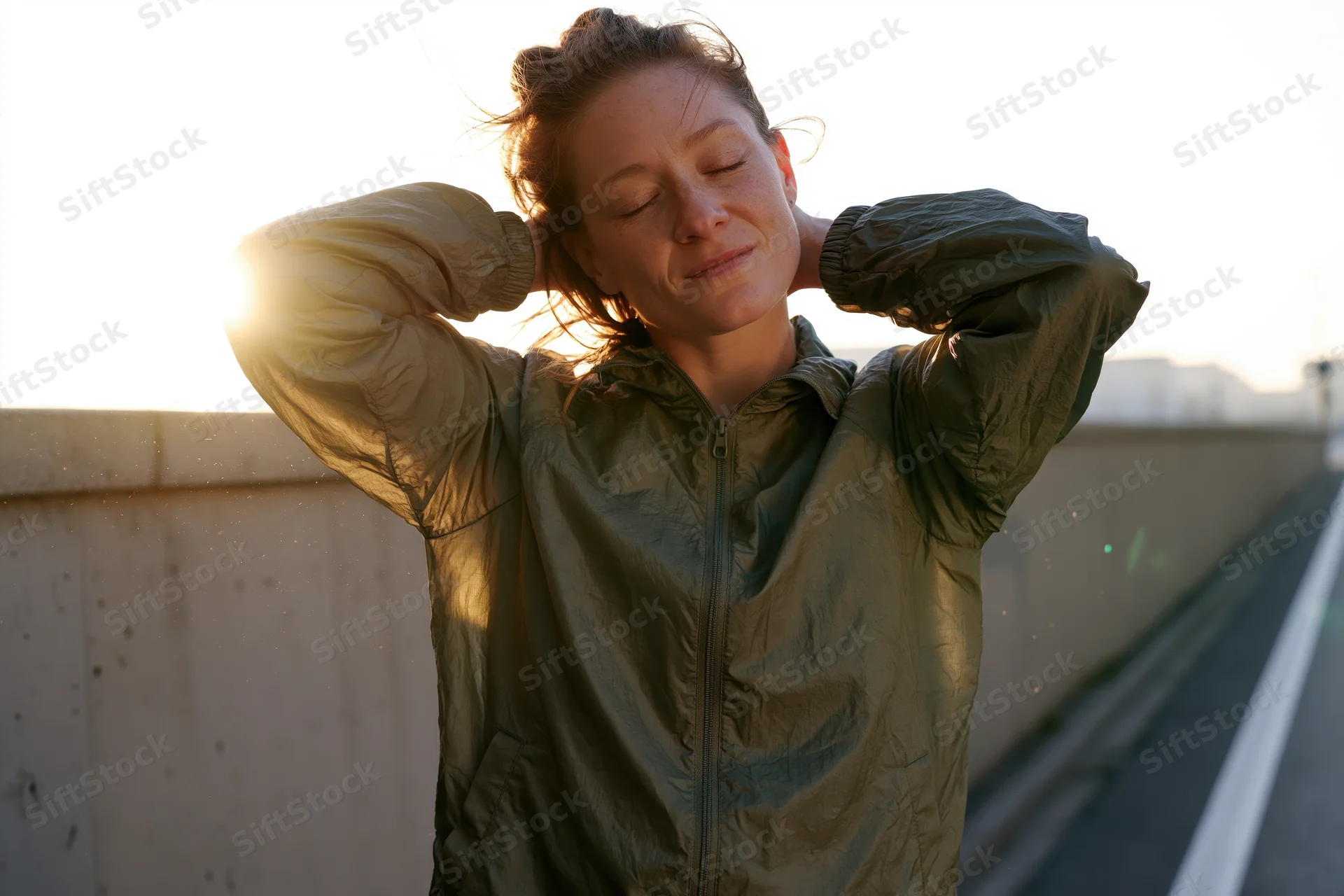 Young woman stretching with hands behind head on sunlit road