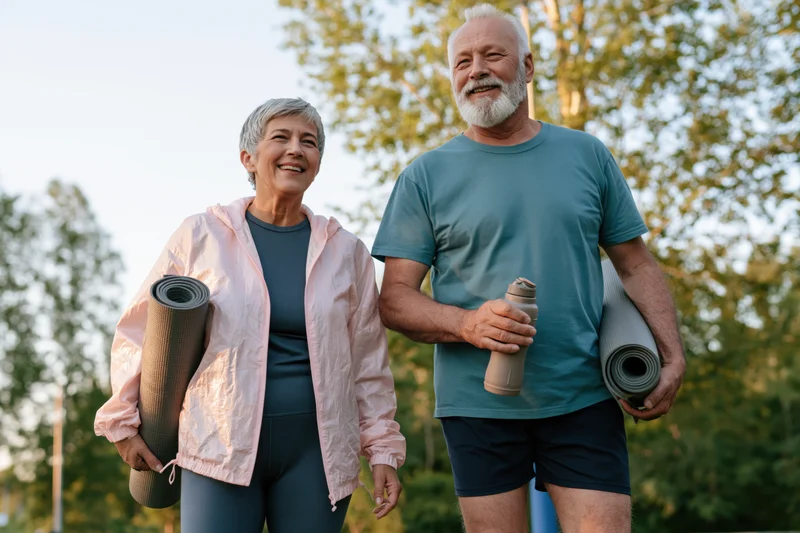 Senior couple smiling while walking outdoors with yoga mats and water bottle in a park