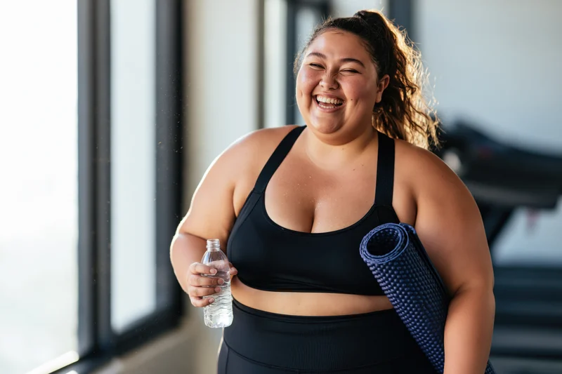 Plus-size woman smiling while holding water bottle and yoga mat in gym