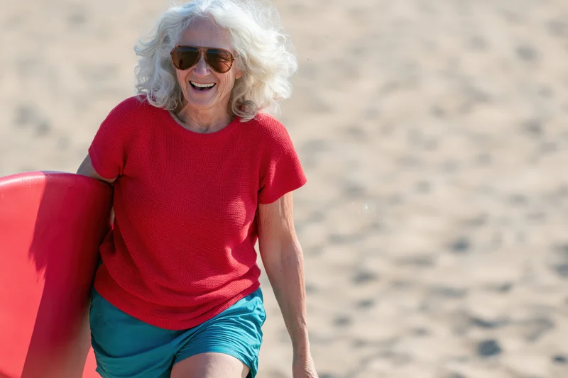 Senior woman smiling while carrying a red surfboard on a sandy beach