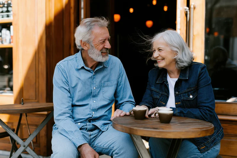 Senior couple smiling and laughing while sitting at a wooden table with coffee cups outside a cafe
