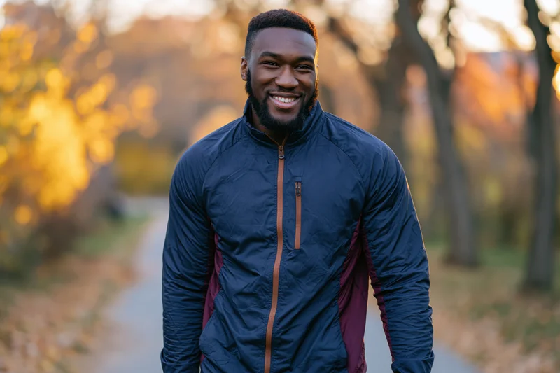 Smiling black man in navy jacket walking on park path during autumn golden hour