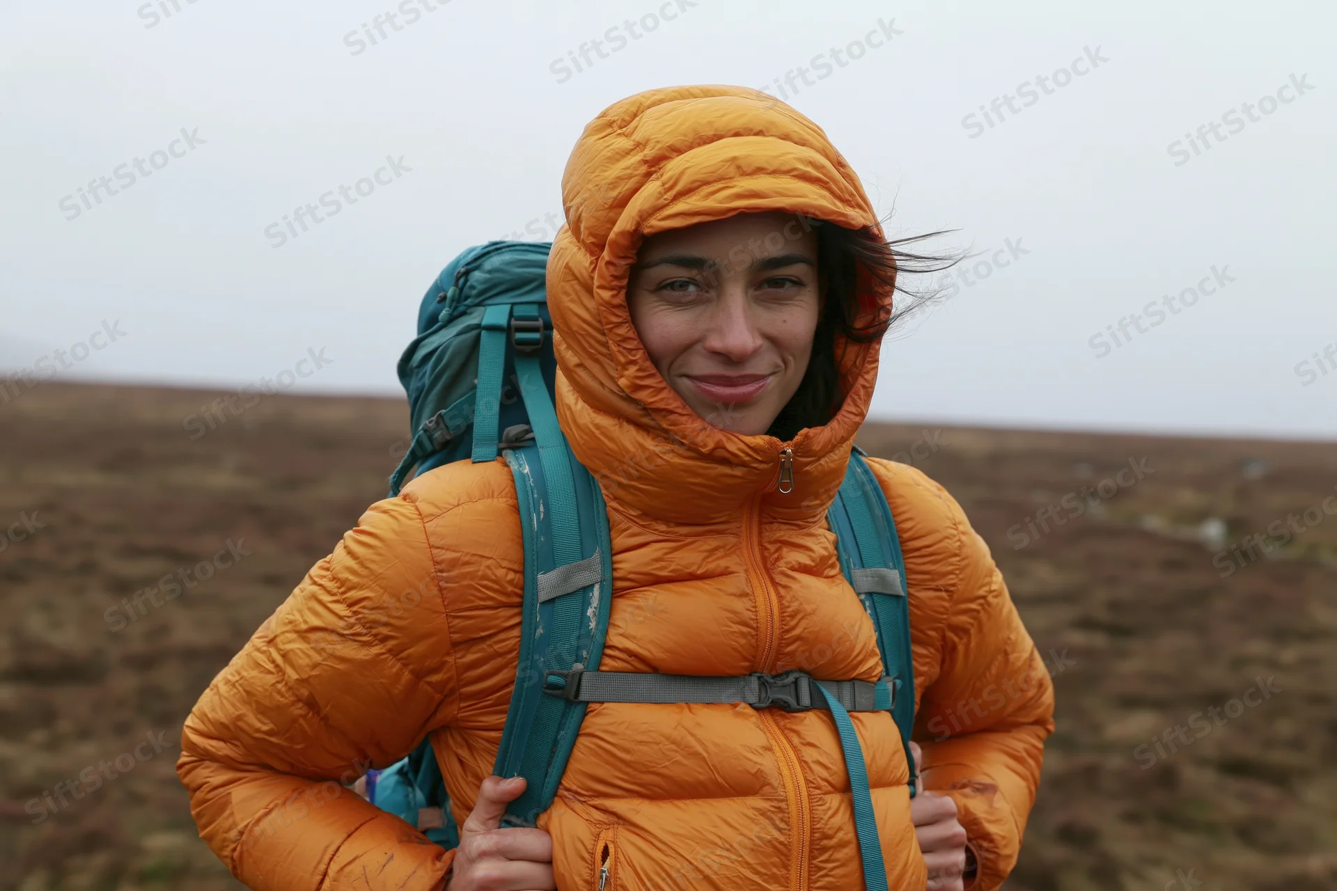 Woman hiking with backpack in moorland landscape, wearing orange hooded jacket, smiling at camera