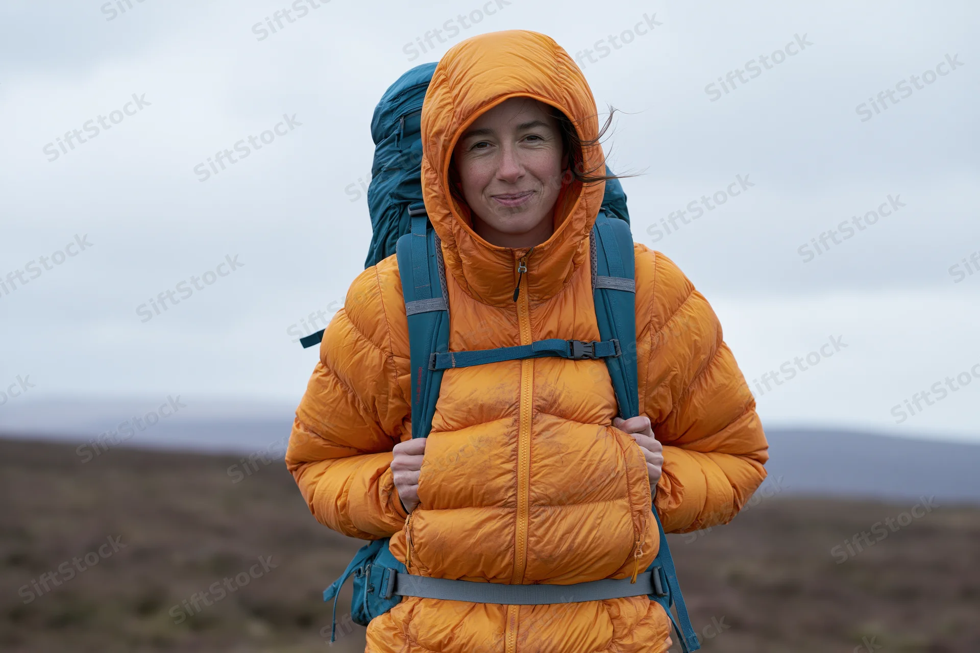 Woman in orange puffer jacket with backpack hiking in overcast moorland