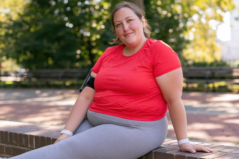 Woman in red shirt and gray leggings sitting on brick bench in park smiling at camera
