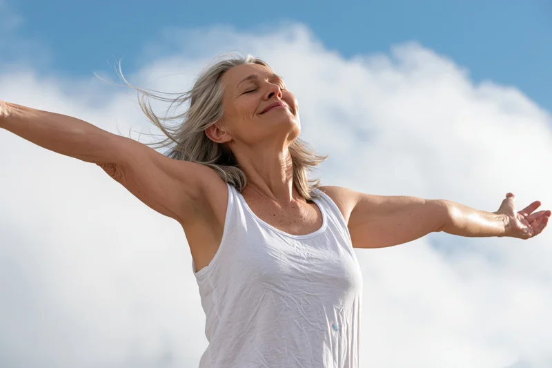 Senior woman with arms outstretched smiling against blue sky and clouds