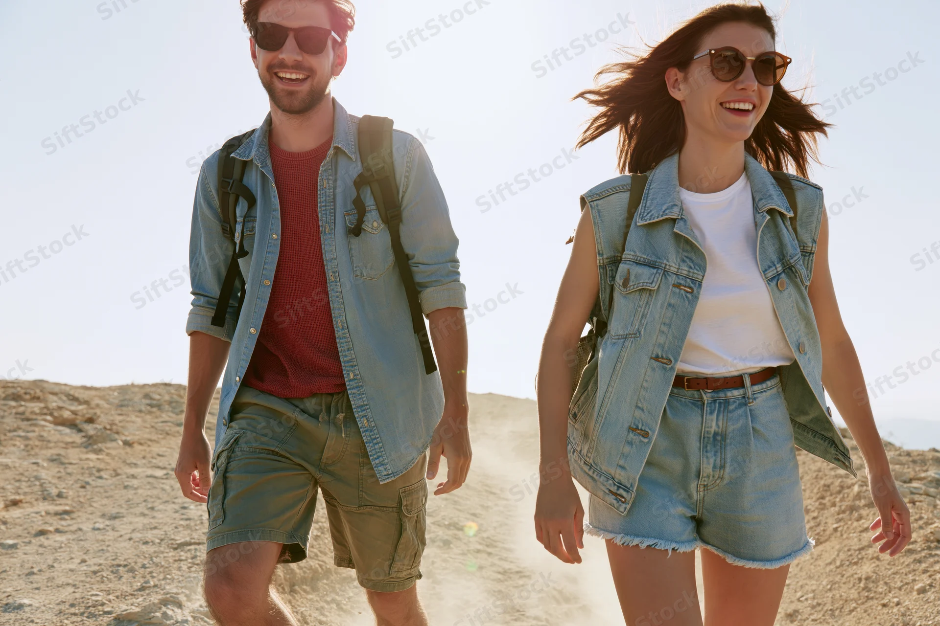 Couple hiking on a sandy trail in daylight, smiling and wearing backpacks and sunglasses