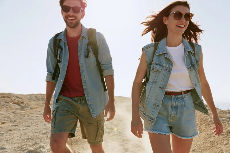 Couple hiking on a sandy trail in daylight, smiling and wearing backpacks and sunglasses