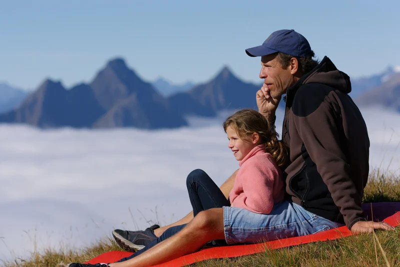 Man and young girl sitting on grassy mountain summit above clouds, overlooking peaks