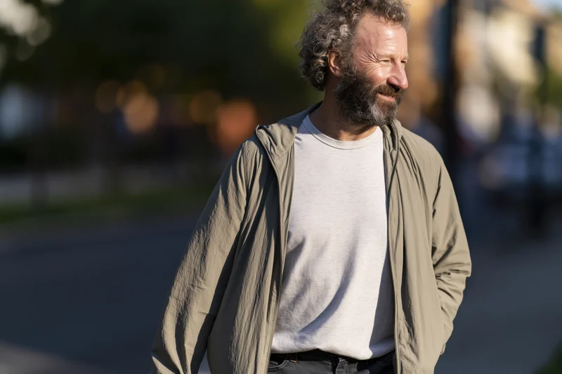 Middle-aged man with curly hair and beard walking outdoors on a city sidewalk, smiling casually in daylight