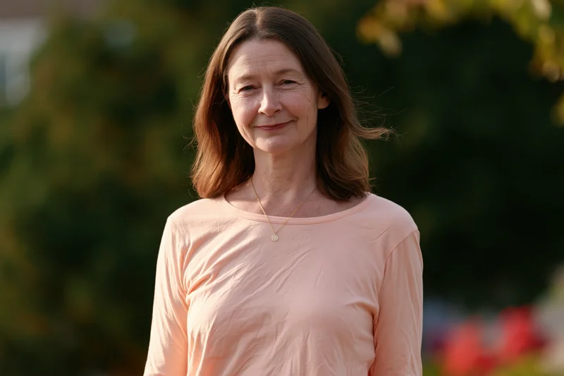 Middle-aged woman smiling outdoors in a garden setting wearing a peach shirt and gold necklace
