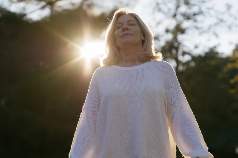 Middle-aged woman with eyes closed standing outdoors in sunlight, surrounded by trees