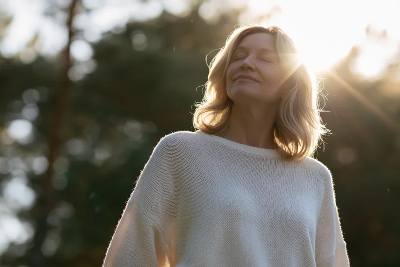 Middle-aged woman smiling with eyes closed in sunlight outdoors among trees