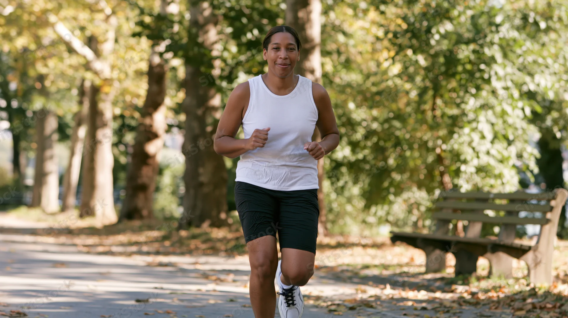 Woman jogging on a paved path in a park with trees and fallen leaves around her