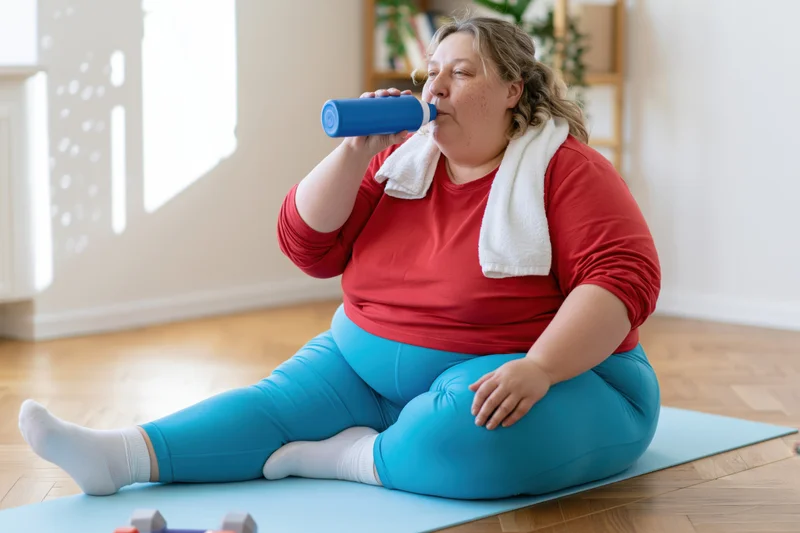 Plus-size woman sitting cross-legged on yoga mat drinking from water bottle after workout at home