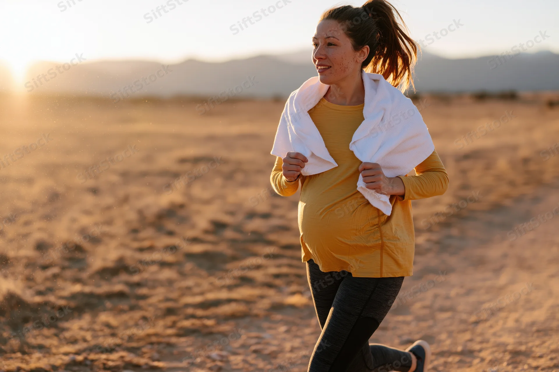 Pregnant woman running outdoors in desert at sunset with towel around neck