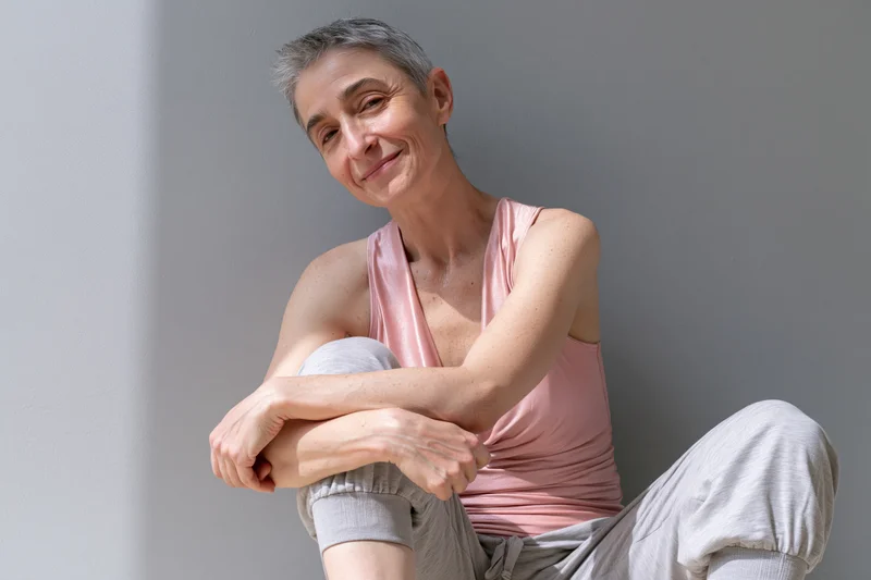 Middle-aged woman smiling while sitting cross-legged against a gray wall in casual clothing