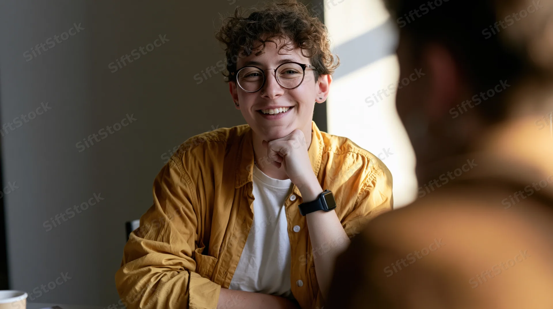 Young woman smiling during a conversation indoors, hand on chin, wearing glasses and a yellow shirt