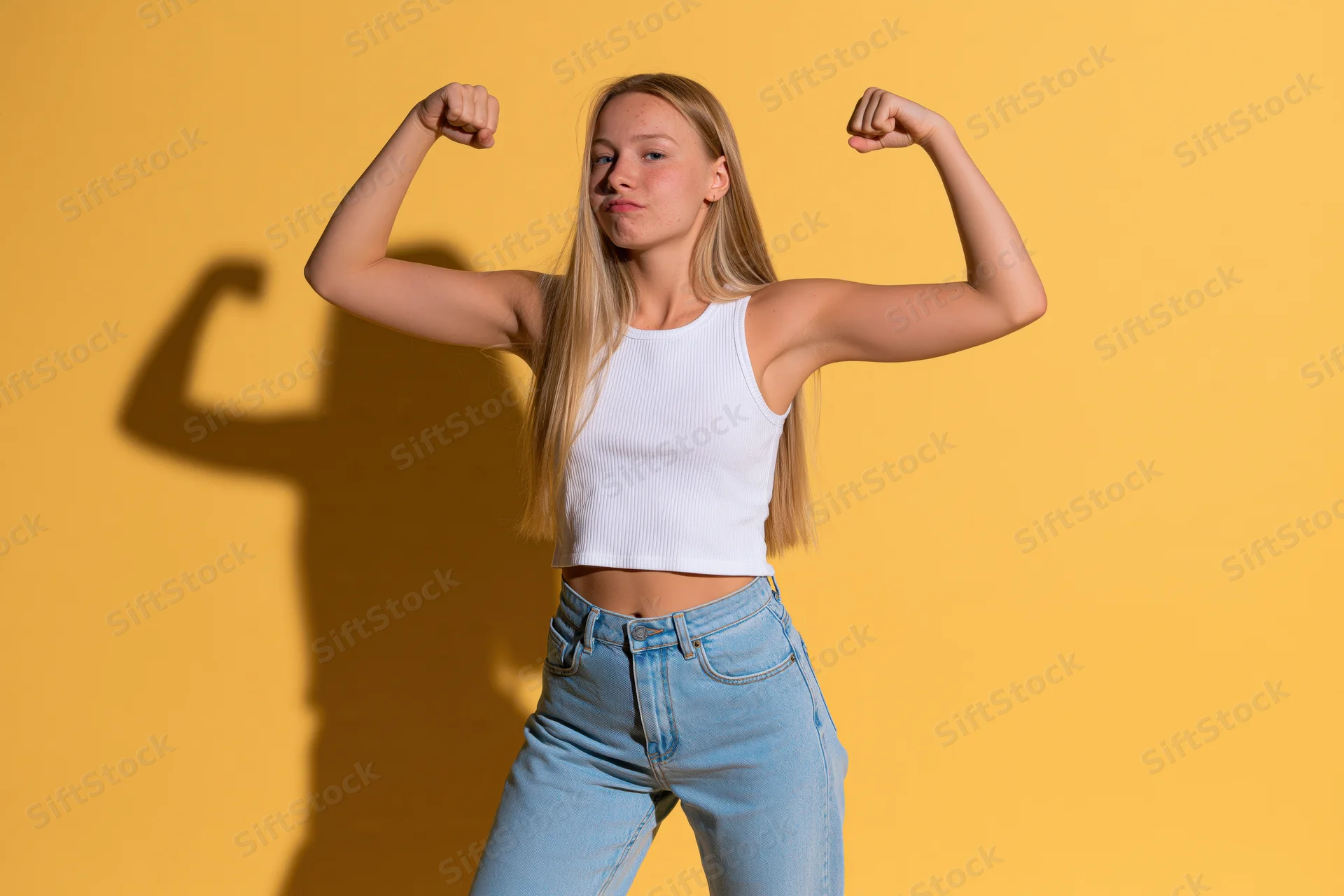 Young woman flexing biceps in white crop top and jeans against yellow background