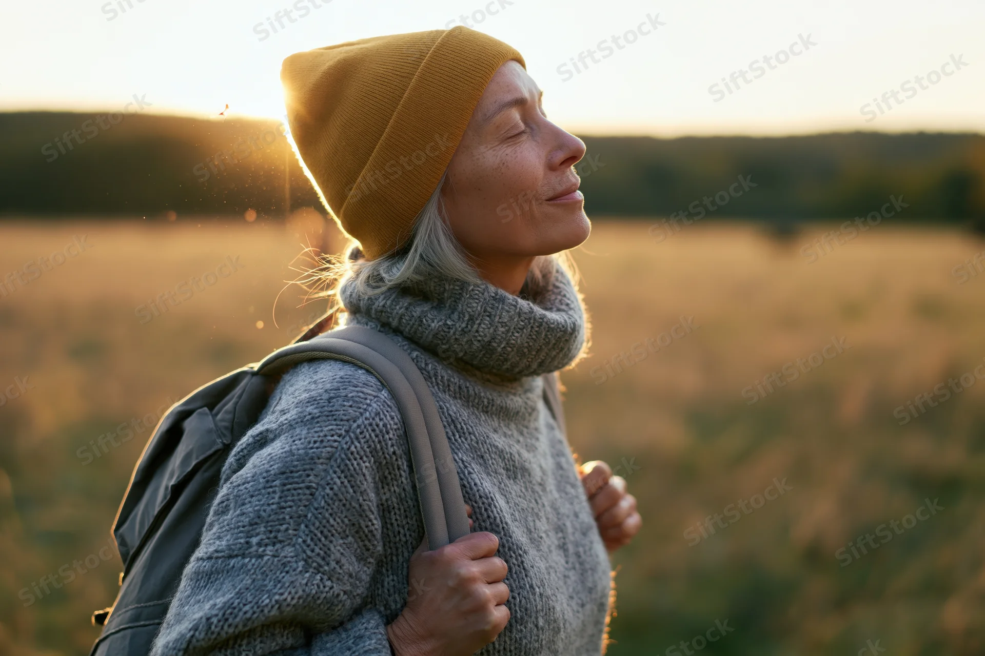 Senior woman with backpack and knitwear standing in a sunlit field at sunset