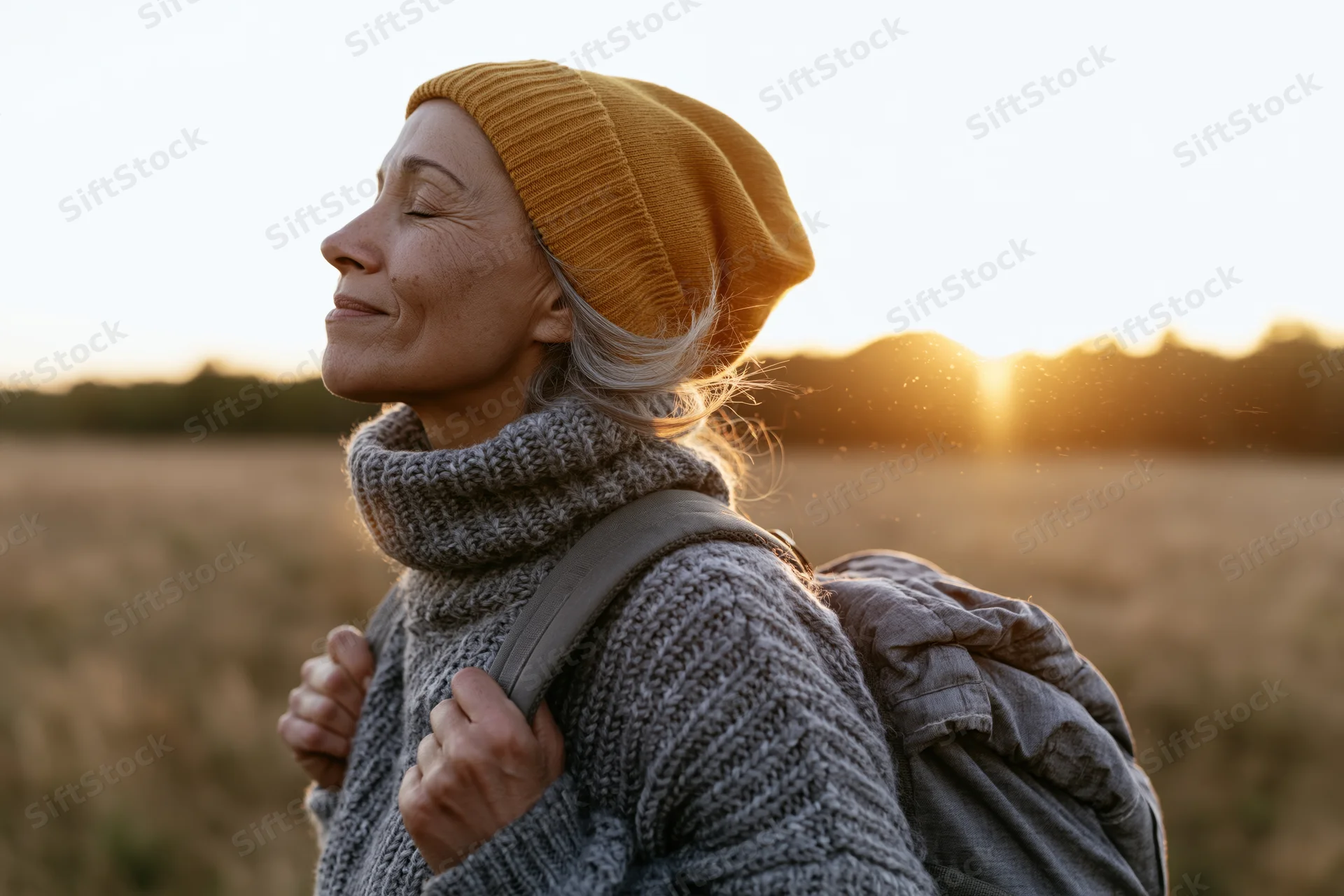 Senior woman with backpack smiling with eyes closed in a sunlit field at sunset