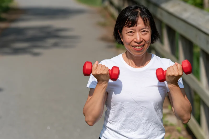 Asian senior woman smiling while lifting red dumbbells outdoors on a paved path