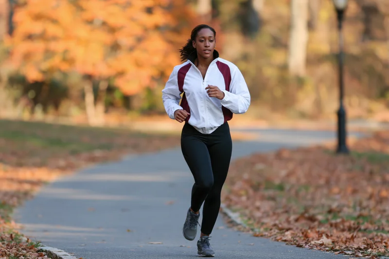 Woman jogging on a winding paved path in a park lined with autumn foliage