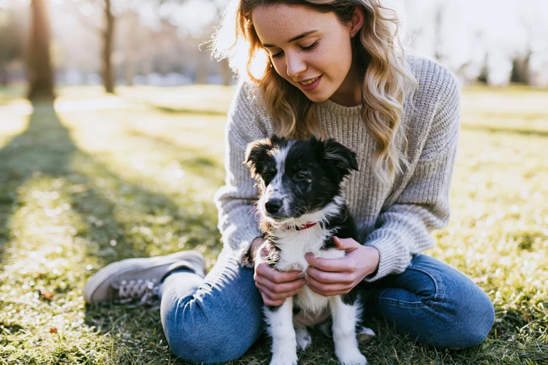 Woman kneeling on grass holding a small black and white puppy in a park