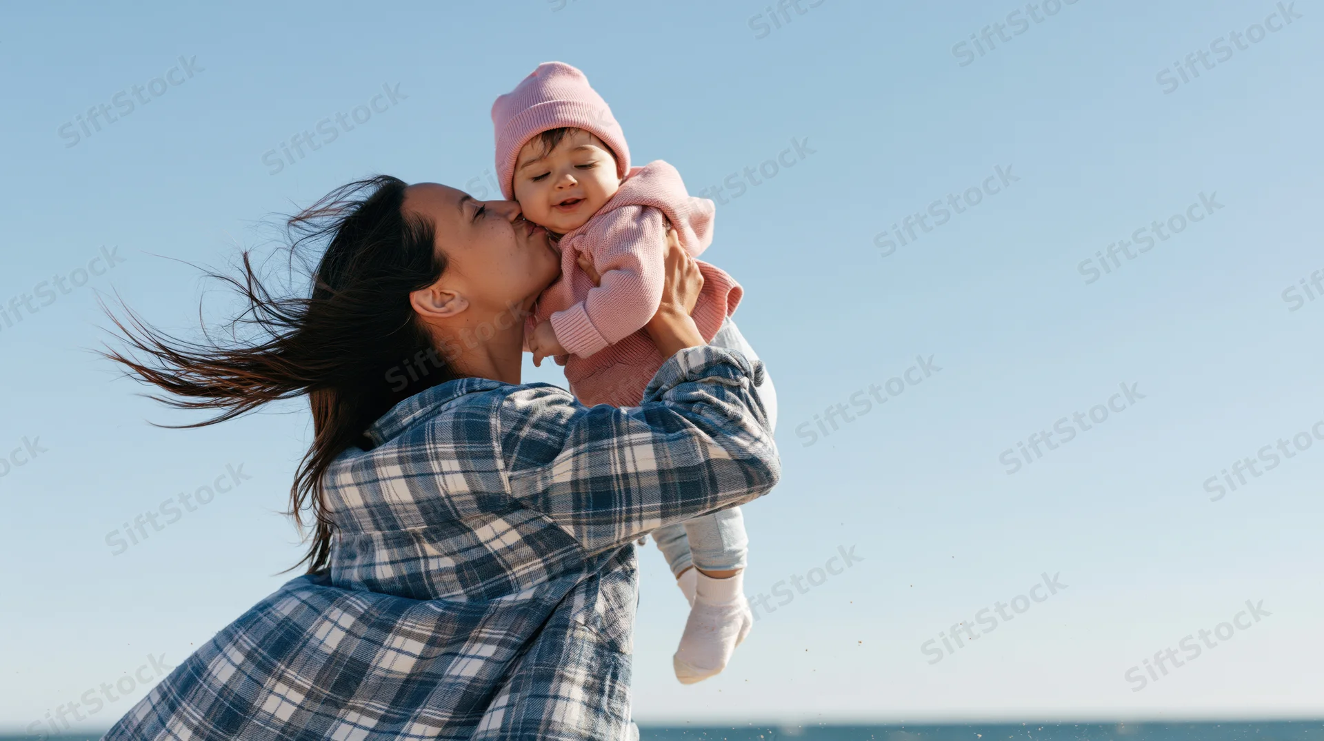 Woman kissing baby while holding her up on a windy beach