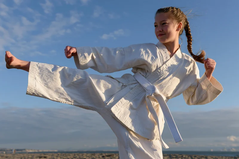 Young girl performing a high kick in karate uniform on a beach under blue sky