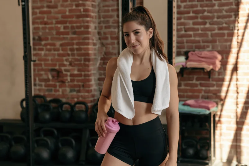 Young woman in gym holding pink water bottle with towel around neck