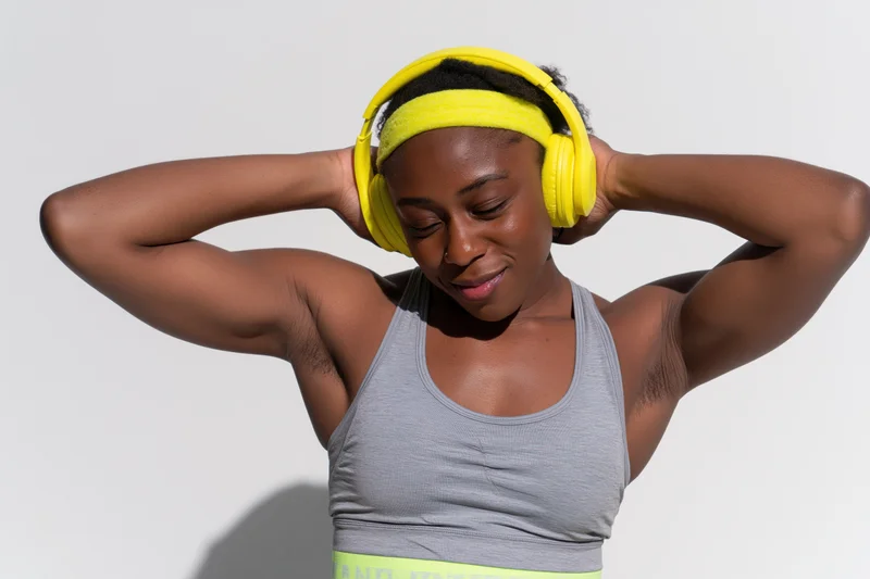 Young black woman smiling with eyes closed while adjusting yellow headphones over her head, wearing gray tank top against white background