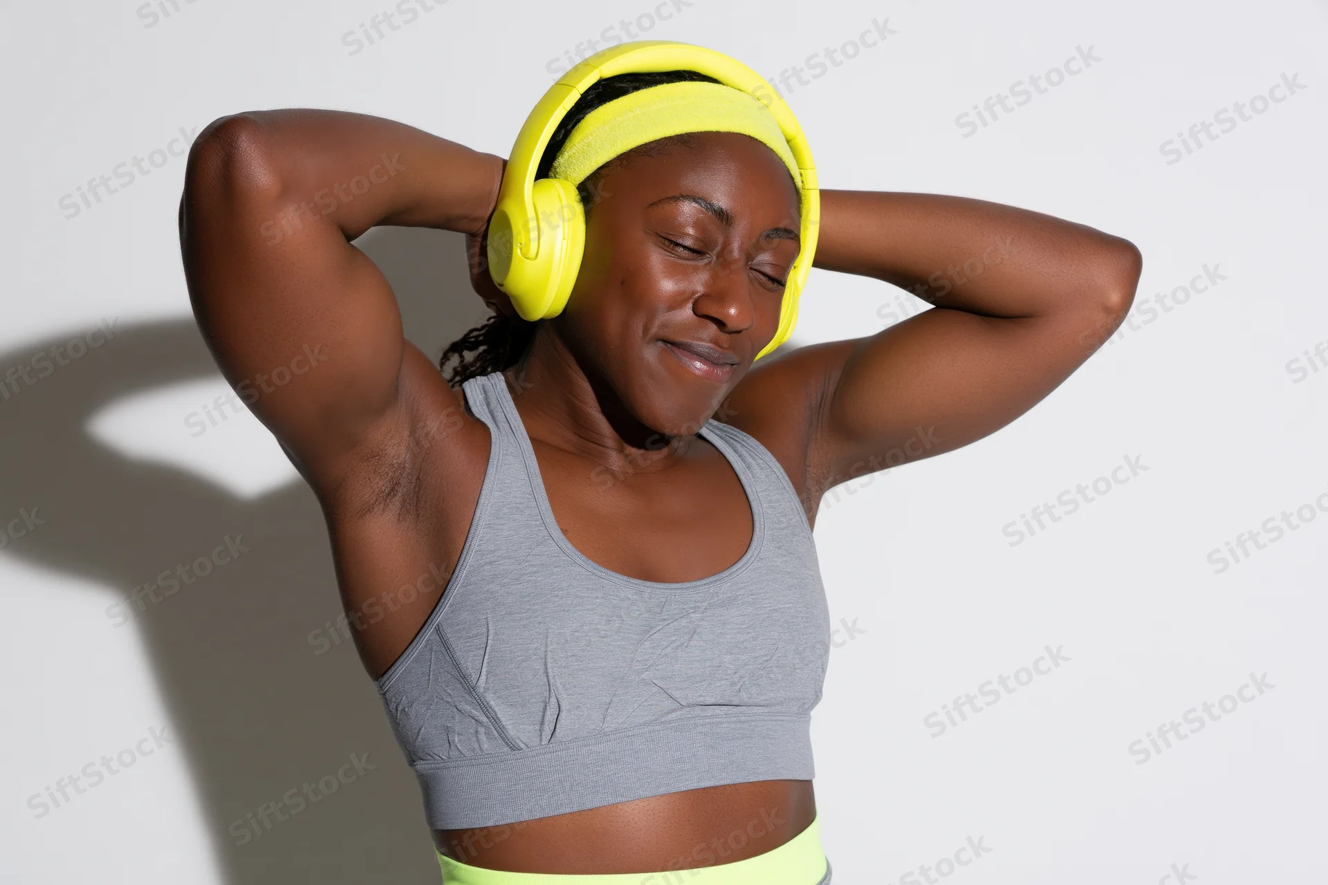 Young black woman smiling with eyes closed, wearing yellow headphones and gray sports bra, arms raised behind head against white background