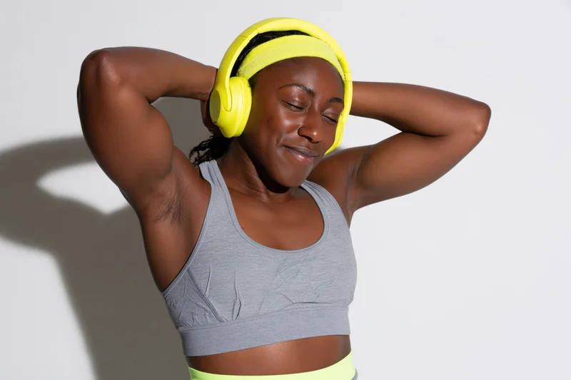 Young black woman smiling with eyes closed, wearing yellow headphones and gray sports bra, arms raised behind head against white background