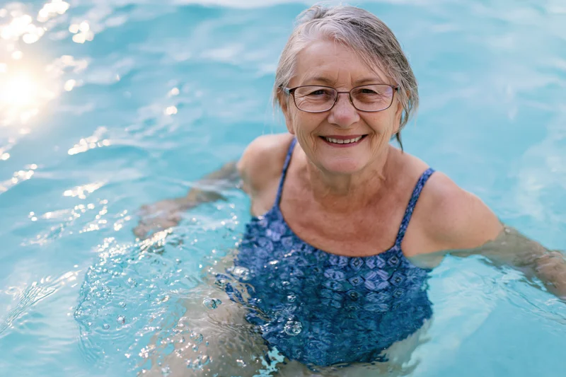 Senior woman smiling while swimming in a pool wearing glasses and blue patterned swimwear