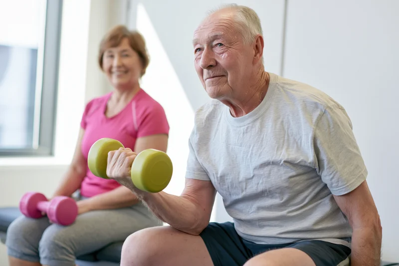 Senior man lifting green dumbbell while seated, woman in pink shirt exercising beside him in bright gym