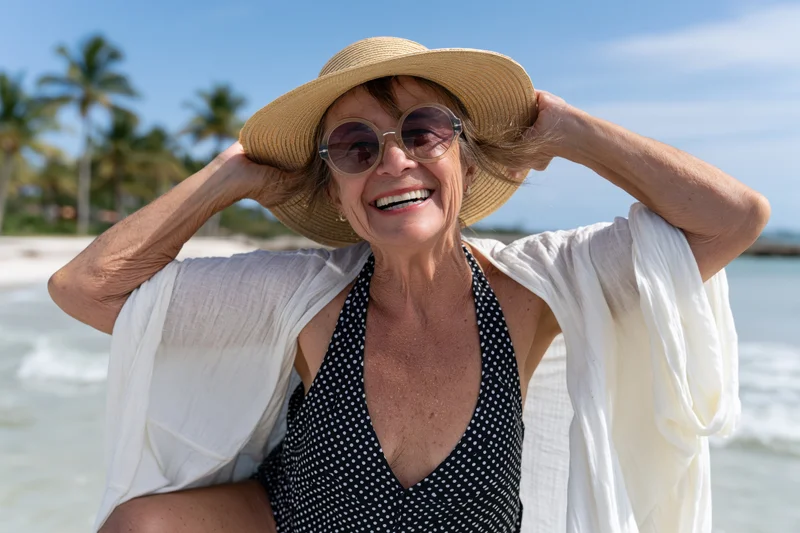Senior woman smiling at beach with straw hat and sunglasses, wearing polka dot swimwear and white coverup, ocean and palm trees in background