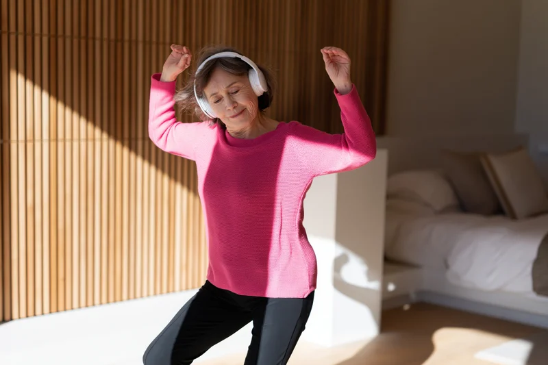Senior woman dancing with headphones in a sunlit bedroom