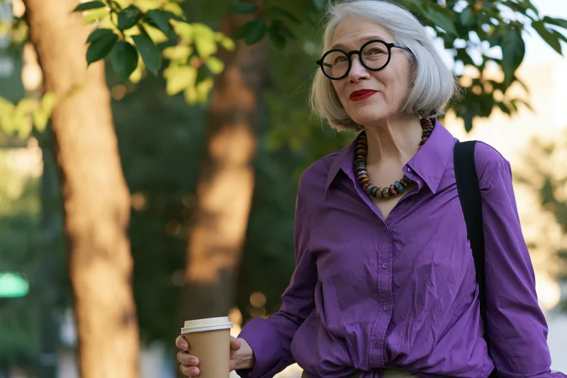 Senior woman holding coffee cup outdoors in park wearing purple shirt and round glasses