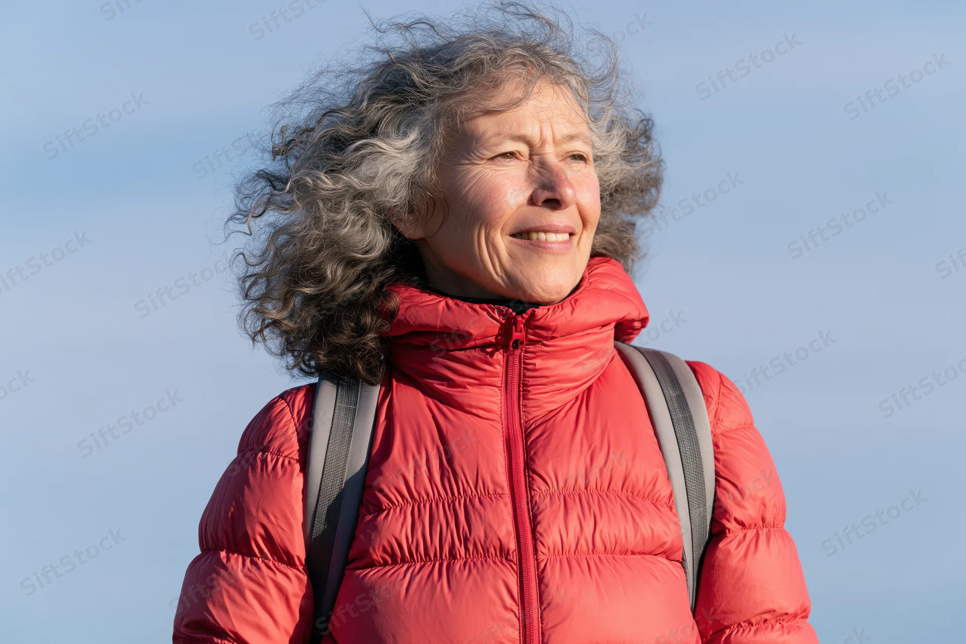 Senior woman smiling outdoors in red puffer jacket with backpack against blue sky