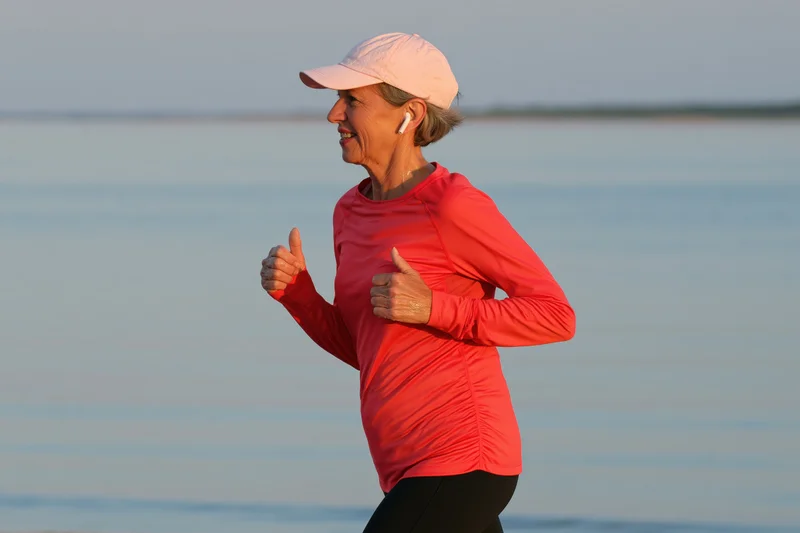 Senior woman running along waterfront in sportswear with earbuds and cap, smiling in morning light