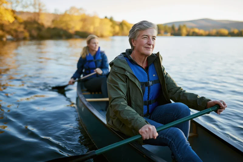 Two women paddling a canoe on a calm lake at sunset surrounded by autumn trees