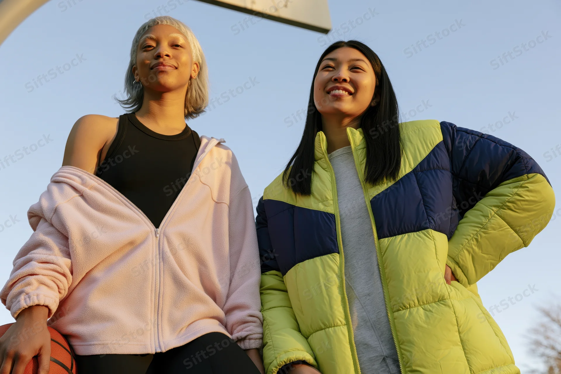 Two women smiling under a basketball hoop outdoors during golden hour