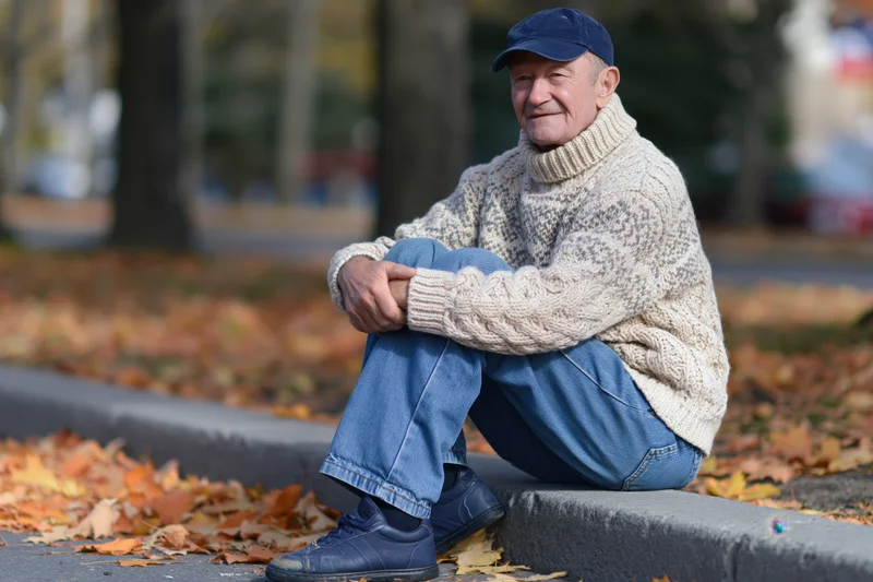 Senior man sitting on curb in autumn park smiling casually