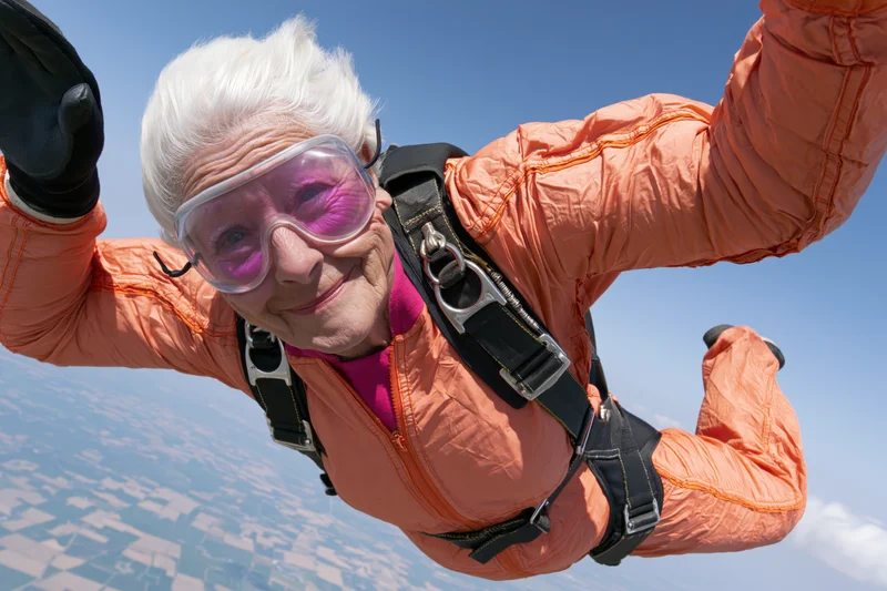 Senior woman skydiving in freefall with orange jumpsuit and goggles, smiling above landscape