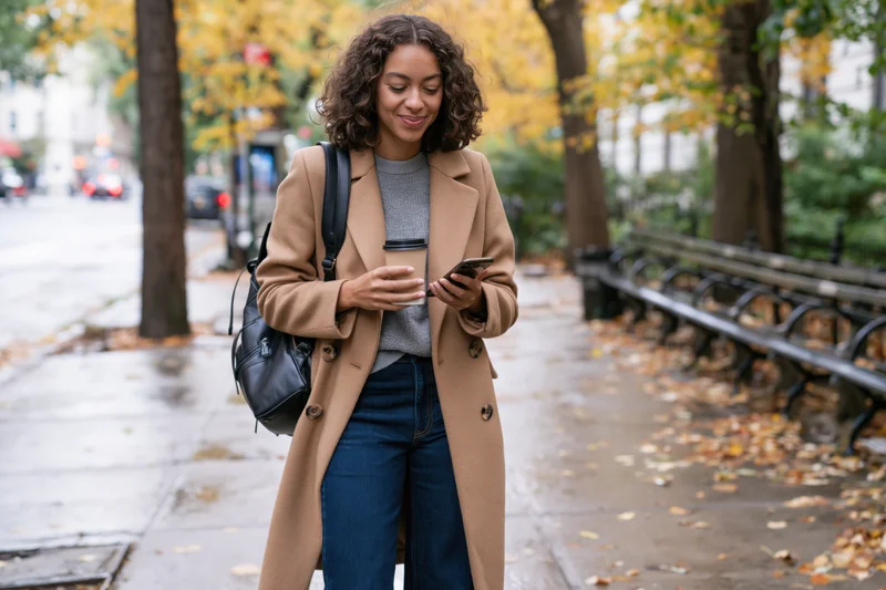 Young black woman walking on sidewalk in autumn park holding smartphone and coffee cup