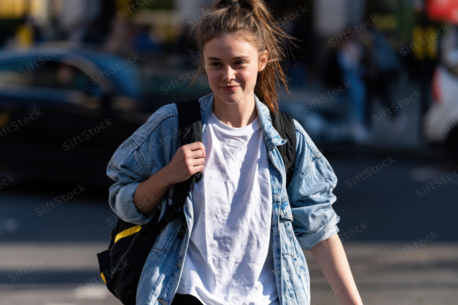 Young woman walking on city sidewalk with backpack, wearing denim jacket and white tshirt