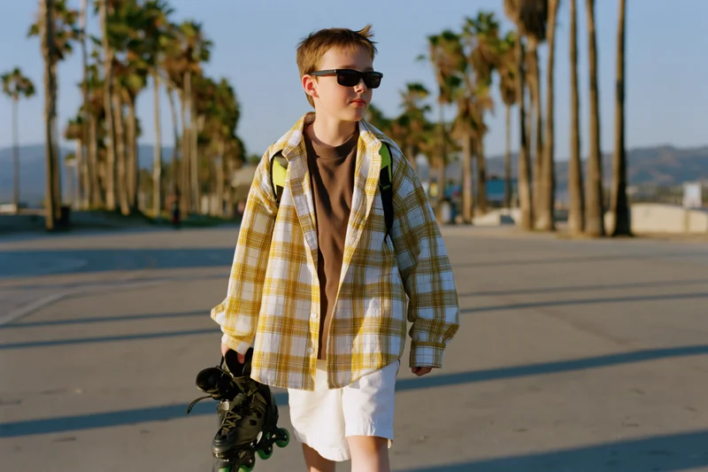 Young boy walking on sidewalk holding roller skates wearing sunglasses and plaid shirt