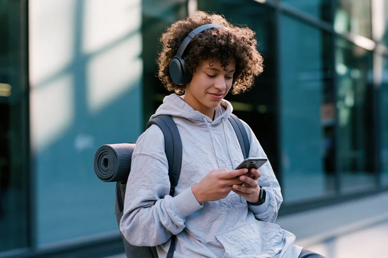 Young person with curly hair wearing headphones and hoodie, sitting outdoors with backpack and yoga mat, using smartphone on city street