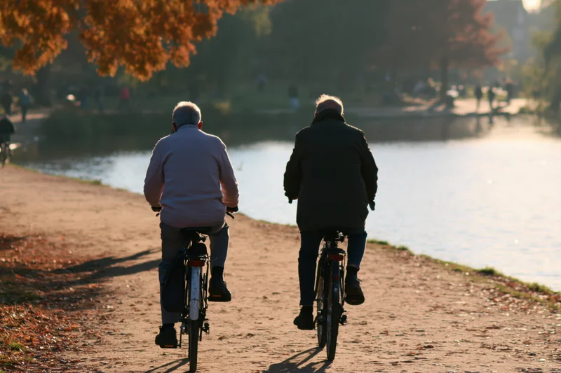 Two elderly men riding bicycles along a lakeside path in autumn